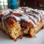 A close-up, low-angle shot of a square piece of moist cinnamon roll casserole topped with white icing.