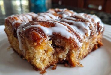 A close-up, low-angle shot of a square piece of moist cinnamon roll casserole topped with white icing.