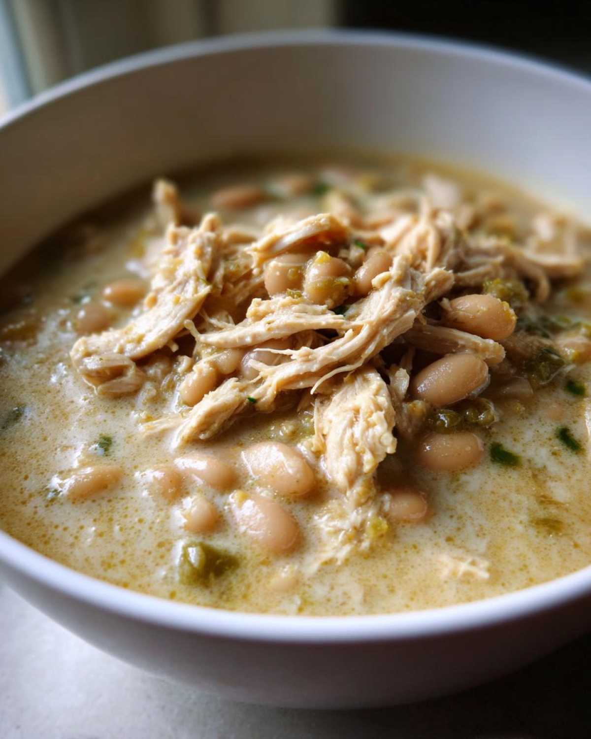 A close-up shot of a white bowl filled with creamy crockpot white chicken chili, topped with shredded chicken and white beans.