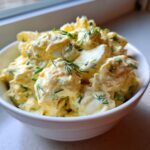 A close-up of a creamy, yellow egg salad recipe garnished with fresh dill and chives in a white bowl.