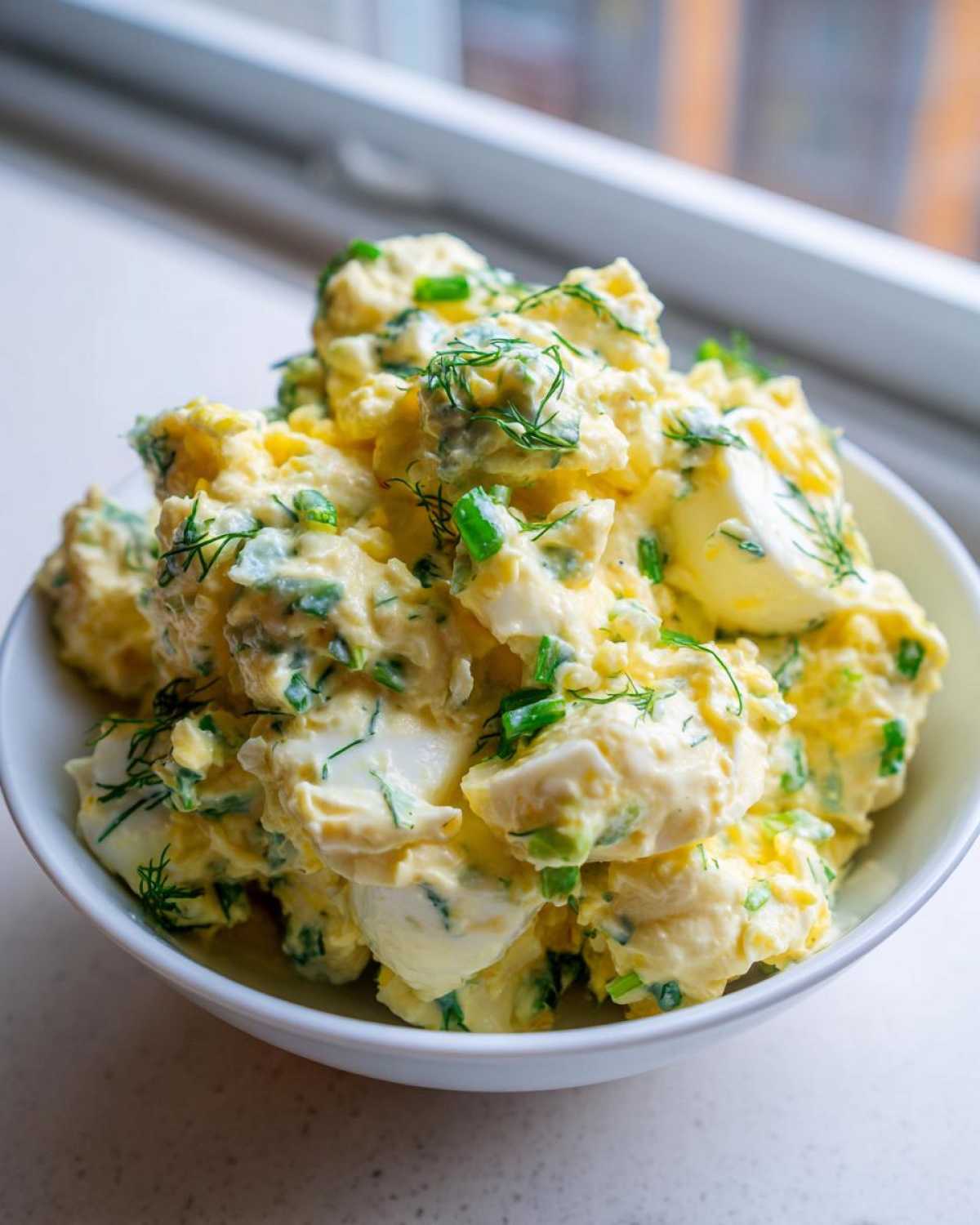 Close-up of creamy egg salad recipe mixed with green onions and dill, served in a white bowl.