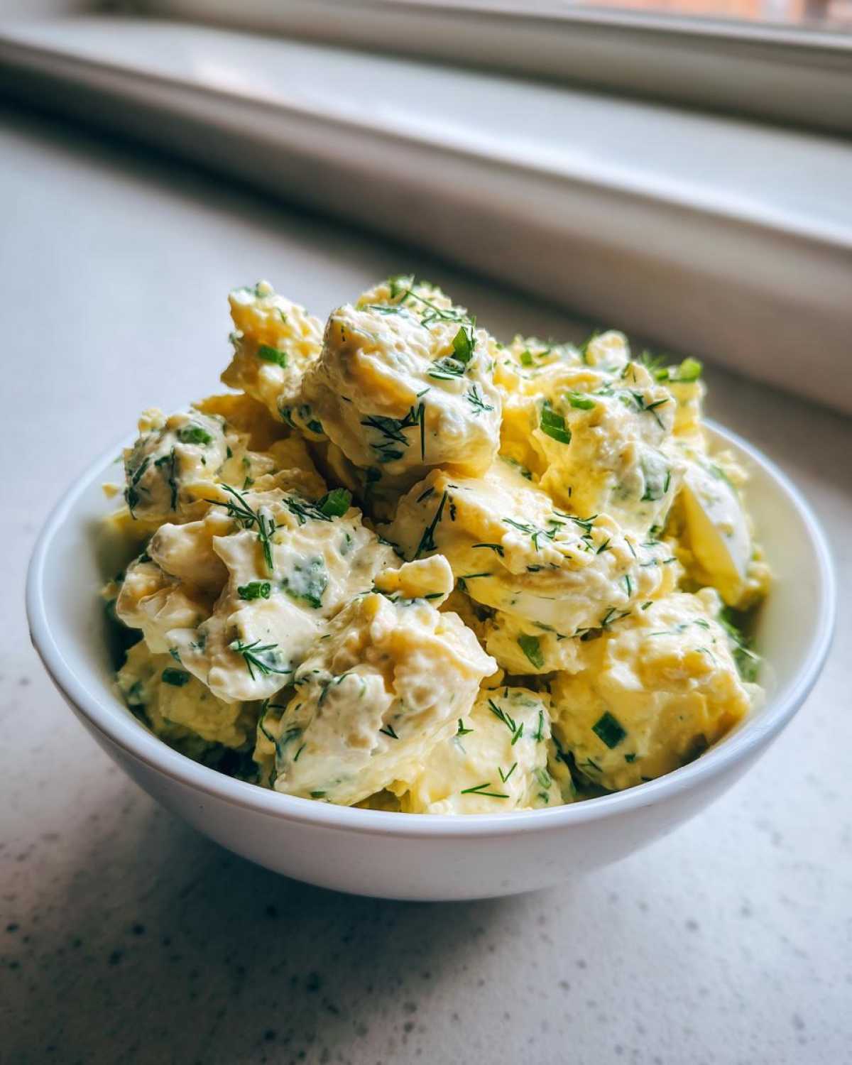 A close-up of creamy egg salad recipe chunks garnished with fresh dill and chives in a white bowl.