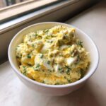 A close-up of creamy, chunky egg salad recipe mixed with fresh dill and chives, served in a white bowl.