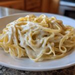A close-up of fettuccine pasta generously coated in rich, creamy homemade alfredo sauce on a white plate.