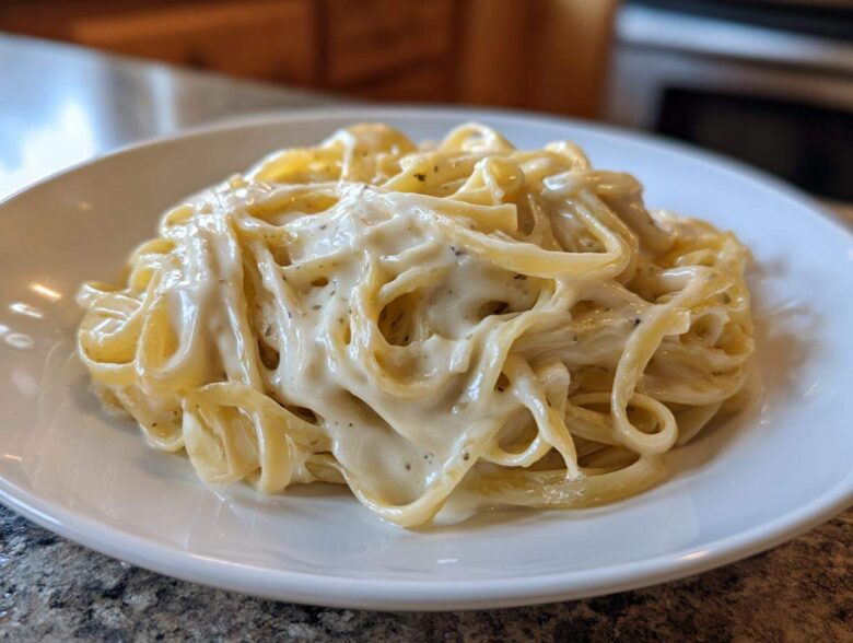 A close-up of fettuccine pasta generously coated in rich, creamy homemade alfredo sauce on a white plate.