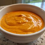 Close-up of a small white bowl filled with thick, vibrant orange nacho cheese sauce.