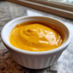 A close-up of thick, vibrant orange nacho cheese sauce swirled in a small white ramekin.