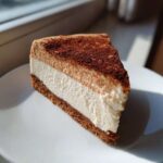 A perfect slice of layered Protein Cheesecake with a graham cracker crust, white filling, and cocoa-dusted top layer.