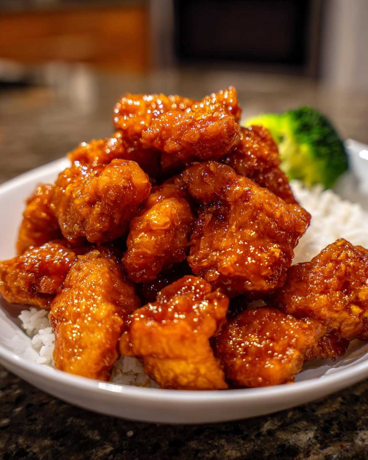 Close-up of crispy orange chicken pieces coated in a glossy sauce, served over white rice with a side of broccoli.