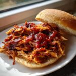 A close-up of a delicious crock pot pulled pork sandwich piled high with saucy meat on a white plate.