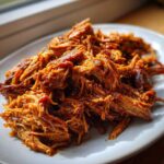 A mound of saucy, shredded crock pot pulled pork piled high on a white plate near a window.