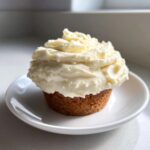 A single cupcake topped high with thick, fluffy cream cheese frosting, sitting on a small white plate.