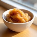Close-up of homemade applesauce, topped with cinnamon, served in a small white bowl near a window.