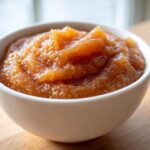 A close-up shot of rich, textured homemade applesauce served in a small white bowl on a wooden surface.