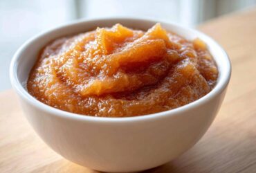 A close-up shot of rich, textured homemade applesauce served in a small white bowl on a wooden surface.