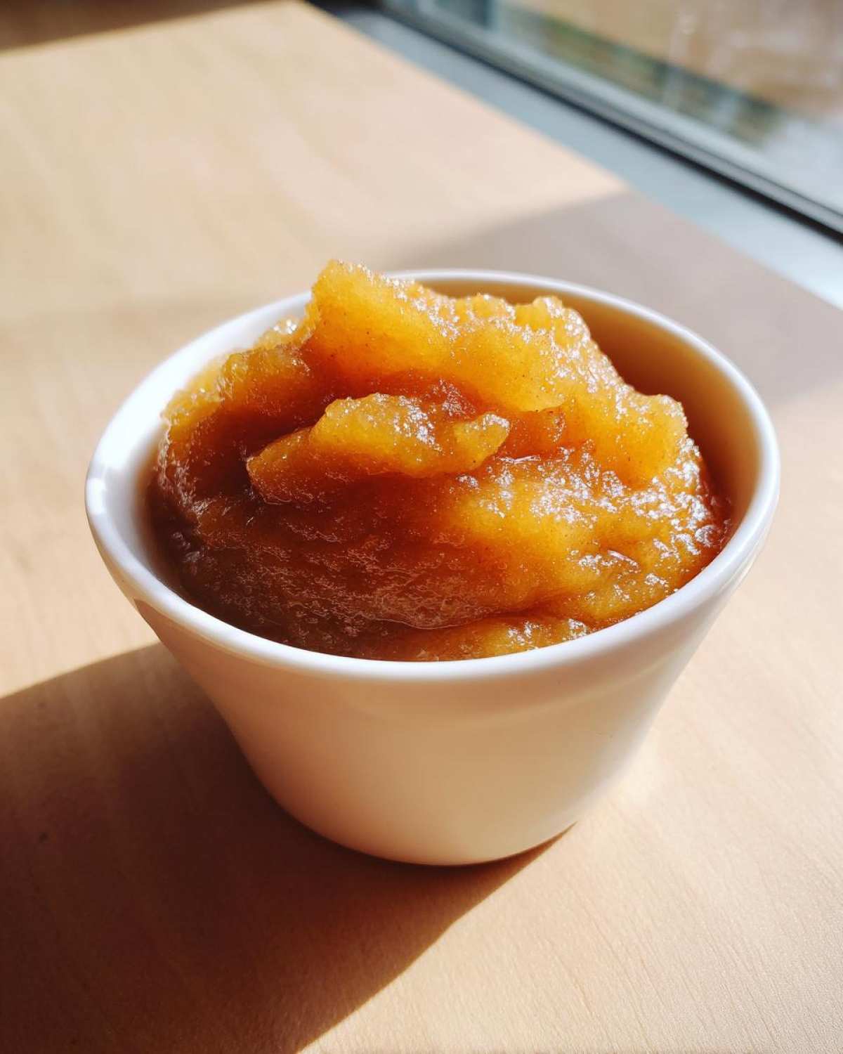 A small white bowl filled with chunky, golden-brown homemade applesauce, highlighted by sunlight.