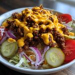 A close-up view of a vibrant cheeseburger bowl featuring seasoned ground beef, melted cheese sauce, pickles, tomatoes, and red onion over lettuce.