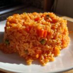 A serving of bright orange Spanish rice, molded into a neat mound on a white plate, showing visible pieces of carrots and peas.