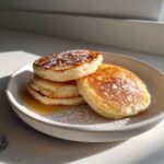 A stack of three fluffy almond flour pancakes dusted with powdered sugar, sitting in a light syrup.