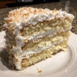 Close-up of a moist slice of three-layer coconut cake with white frosting and toasted coconut flakes.