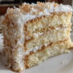 A tall, moist slice of three-layer coconut cake covered in white frosting and toasted coconut flakes.