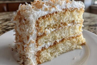 A tall, moist slice of three-layer coconut cake covered in white frosting and toasted coconut flakes.