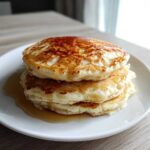 A stack of three fluffy greek yogurt pancakes drizzled generously with golden syrup on a white plate.