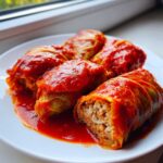 Four savory cabbage rolls covered in rich tomato sauce served on a white plate, one roll is cut open showing rice and meat filling.