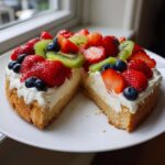 A slice removed from a fruit pizza sugar cookie topped with cream cheese frosting, strawberries, kiwi, and blueberries.