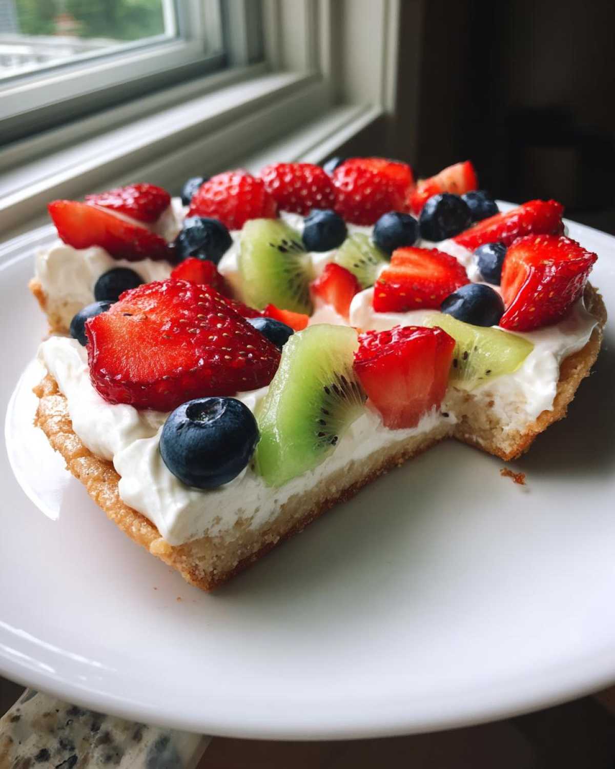 A slice of fruit pizza sugar cookie topped with whipped cream, strawberries, blueberries, and kiwi.