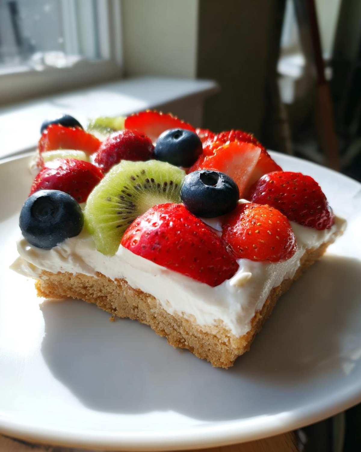 A square slice of fruit pizza sugar cookie topped with cream cheese frosting, strawberries, blueberries, and kiwi.