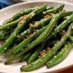 A close-up of bright green beans seasoned with coarse sea salt and browned garlic bits.