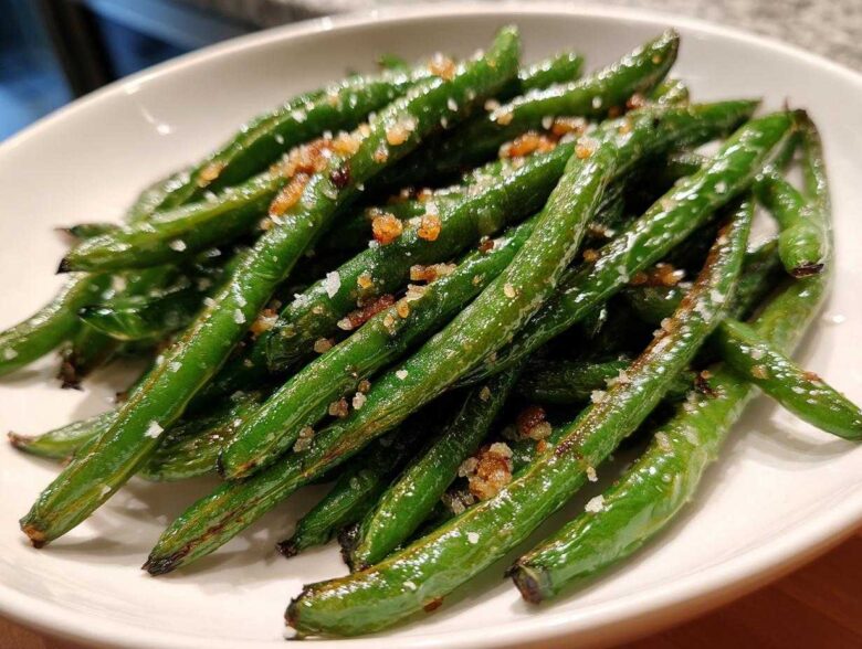 A close-up of bright green beans seasoned with coarse sea salt and browned garlic bits.