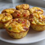 A close-up of six freshly baked Egg Muffin Cups with visible bits of bacon and peppers, served on a white plate.