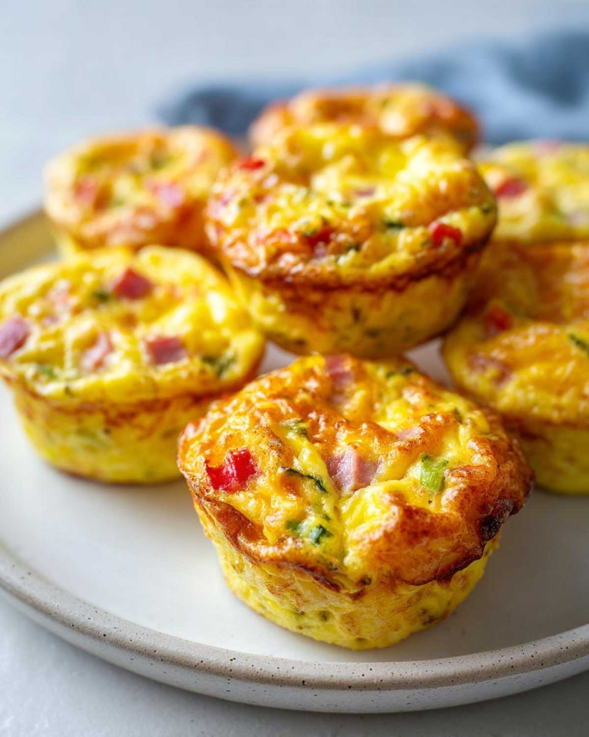 A close-up of several golden brown Egg Muffin Cups filled with visible pieces of ham and red pepper on a light plate.