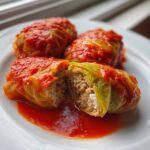Three delicious cabbage rolls served on a white plate, one is cut open showing the ground meat and rice filling.
