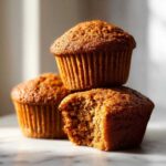 Three golden brown honey bran muffins stacked, with one in the front having a bite taken out to show the moist texture.