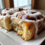 A close-up shot of a fluffy, golden brown cinnamon roll generously drizzled with white icing, with a bite taken out.