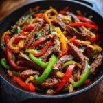 Close-up shot of sizzling steak fajitas with colorful bell peppers and onions in a cast iron skillet.