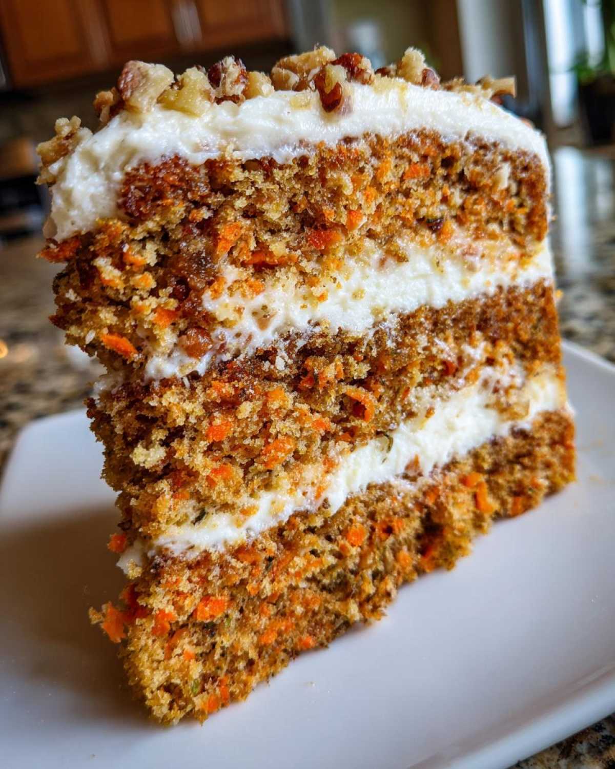 Close-up of a tall, three-layer slice of carrot cake recipe with thick cream cheese frosting and chopped walnuts on top.