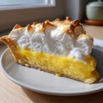 Close-up of a slice of lemon meringue pie showing thick, bright yellow filling and tall, toasted meringue peaks.
