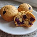 Three blueberry protein muffins on a white plate, one cut open showing the moist interior and berries.