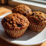 Three freshly baked, moist bran muffins studded with raisins sitting on a white plate in bright sunlight.