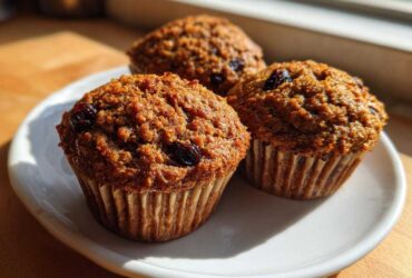 Three freshly baked, moist bran muffins studded with raisins sitting on a white plate in bright sunlight.