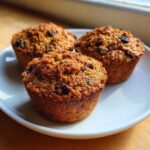 Three golden brown, moist bran muffins studded with raisins sitting on a white plate.