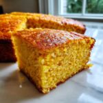 A close-up, appetizing slice of golden buttermilk cornbread resting on a white marble surface.