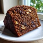 Close-up of a moist slice of date nut bread studded with walnuts and dark raisins, resting on a white plate.