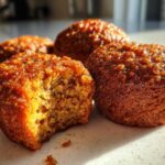 Close-up of several moist, golden-brown honey bran muffins, one with a bite taken out showing the texture.