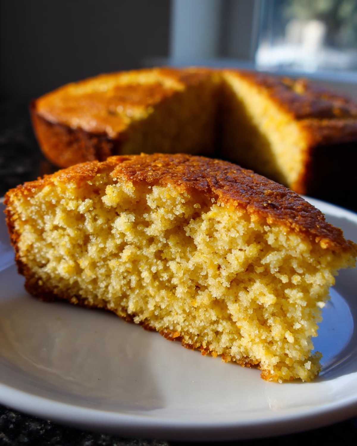 Close-up of a golden, moist slice of skillet cornbread served on a white plate, with the rest of the cornbread visible in the background.