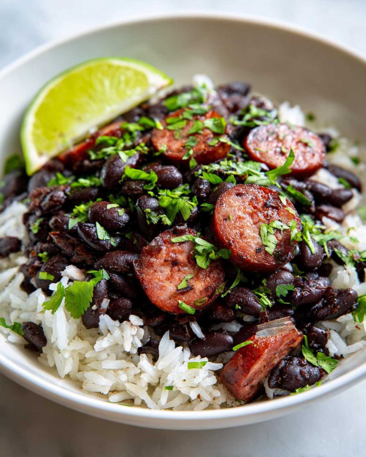 Close-up of a bowl filled with white rice topped with seasoned black beans and rice, sliced smoked sausage, and fresh cilantro, garnished with a lime wedge.